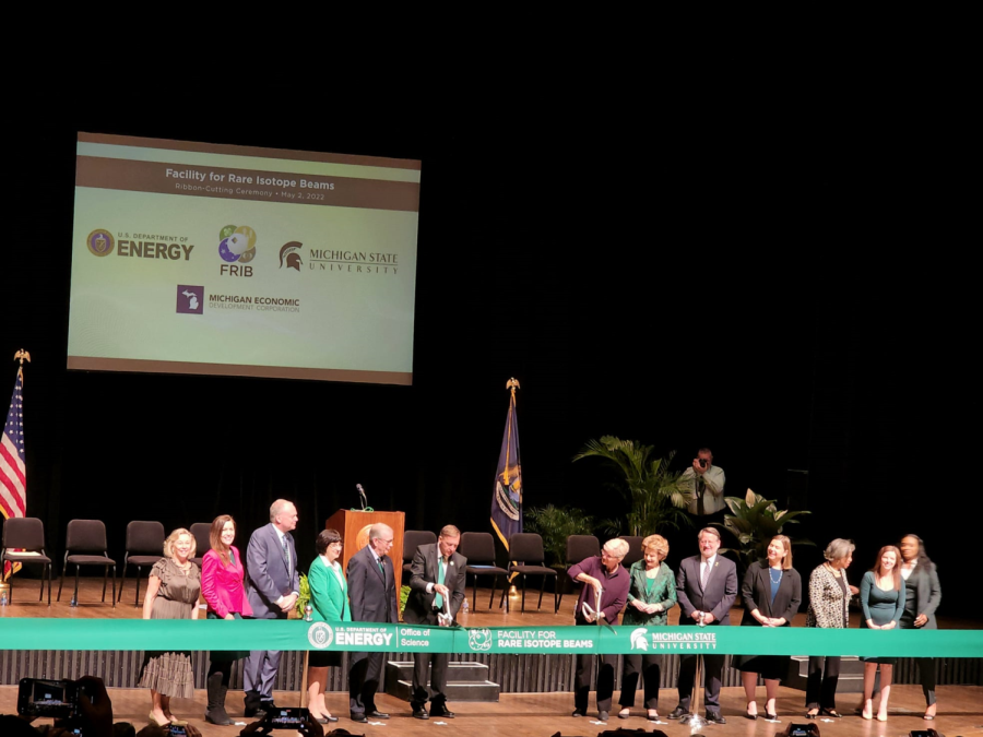 People standing on a stage behind a green ribbon. Two people have large scissors to cut the ribbon, marking the beginning of FRIB.
