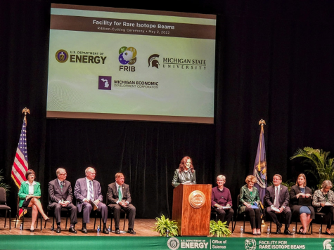 Whitmer stands on a platform on stage with leaders in Michigan sitting behind her. A green ribbon is in front of her.