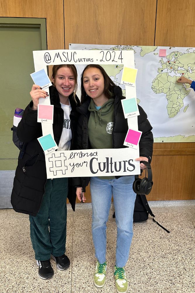 Two students stand in a picture frame prop for the #embraceyourculturs social media campaign.